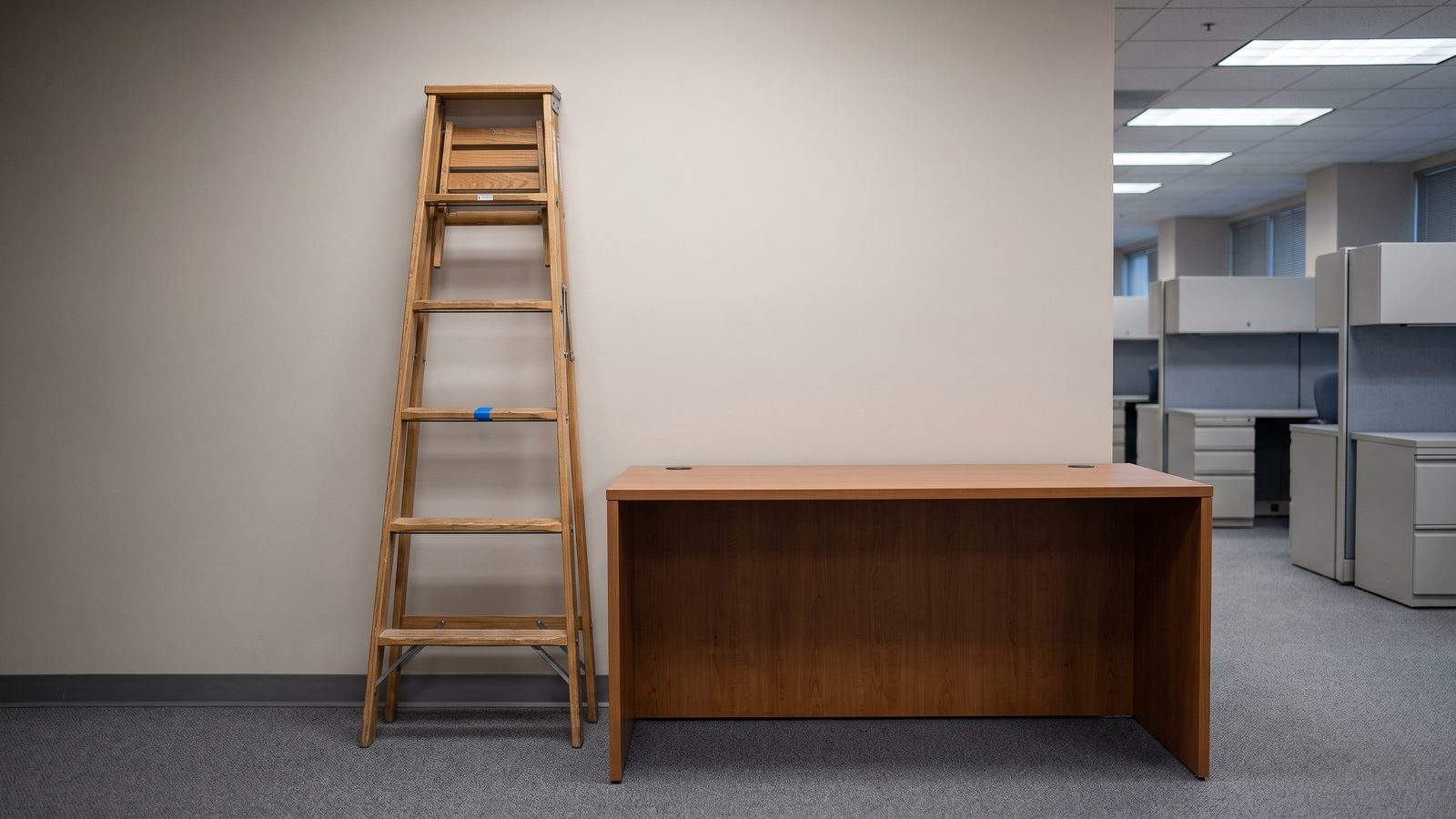A wooden step ladder leaning against a beige office wall beside an empty desk, one rung marked with blue tape