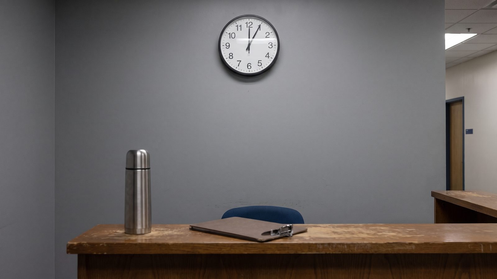 A wall clock above an empty night-shift desk with a thermos and a face-down clipboard