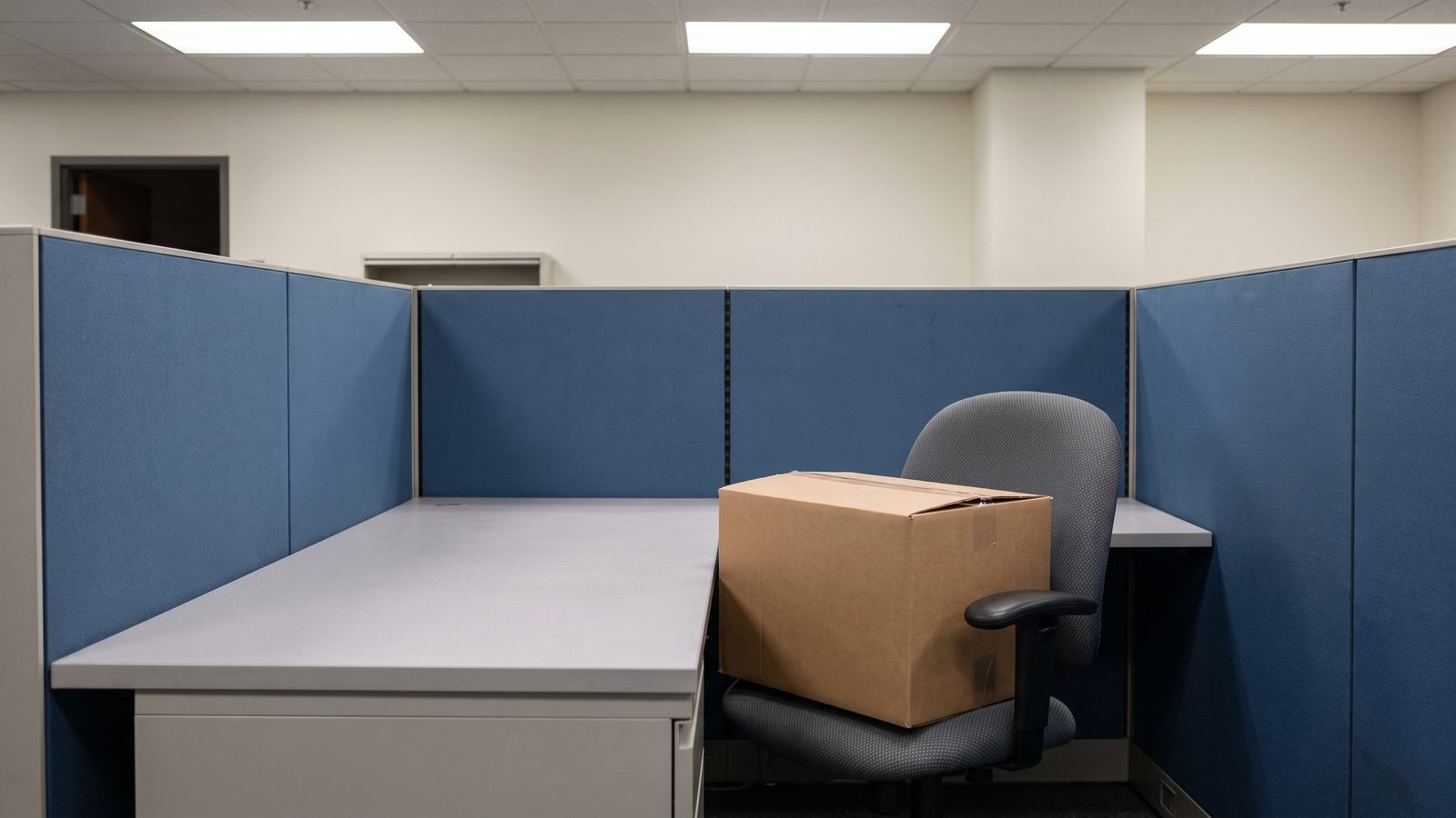 A taped cardboard box on the chair of an empty office cubicle with blue fabric partitions and a bare desk