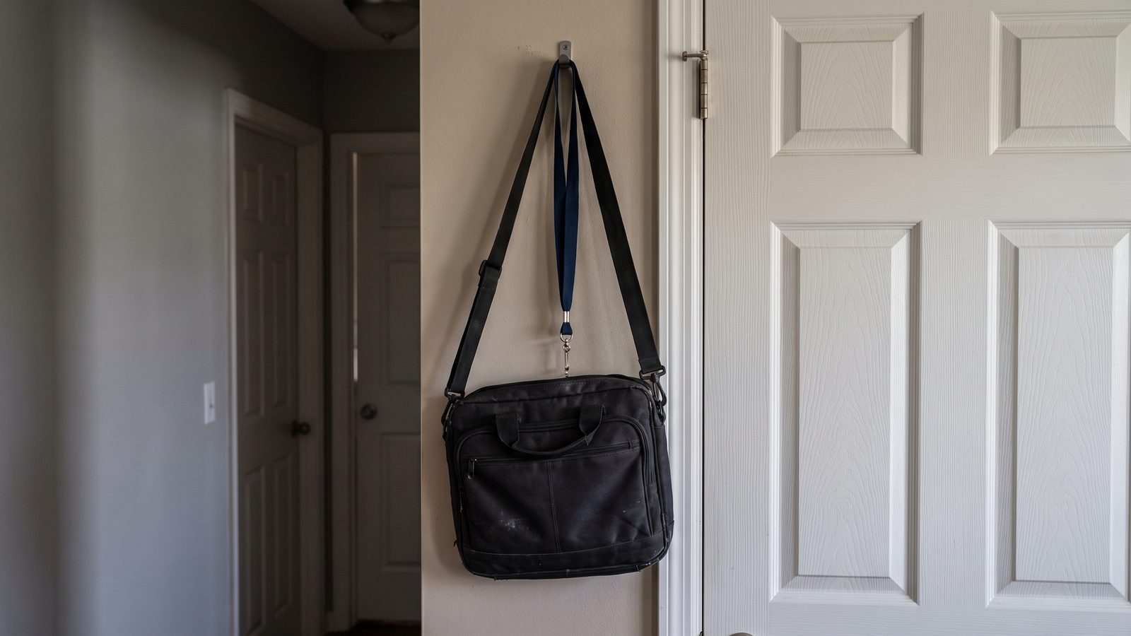 A black laptop bag and a blue lanyard hanging from a single hook on a hallway wall inside a house, beside a closed white front door