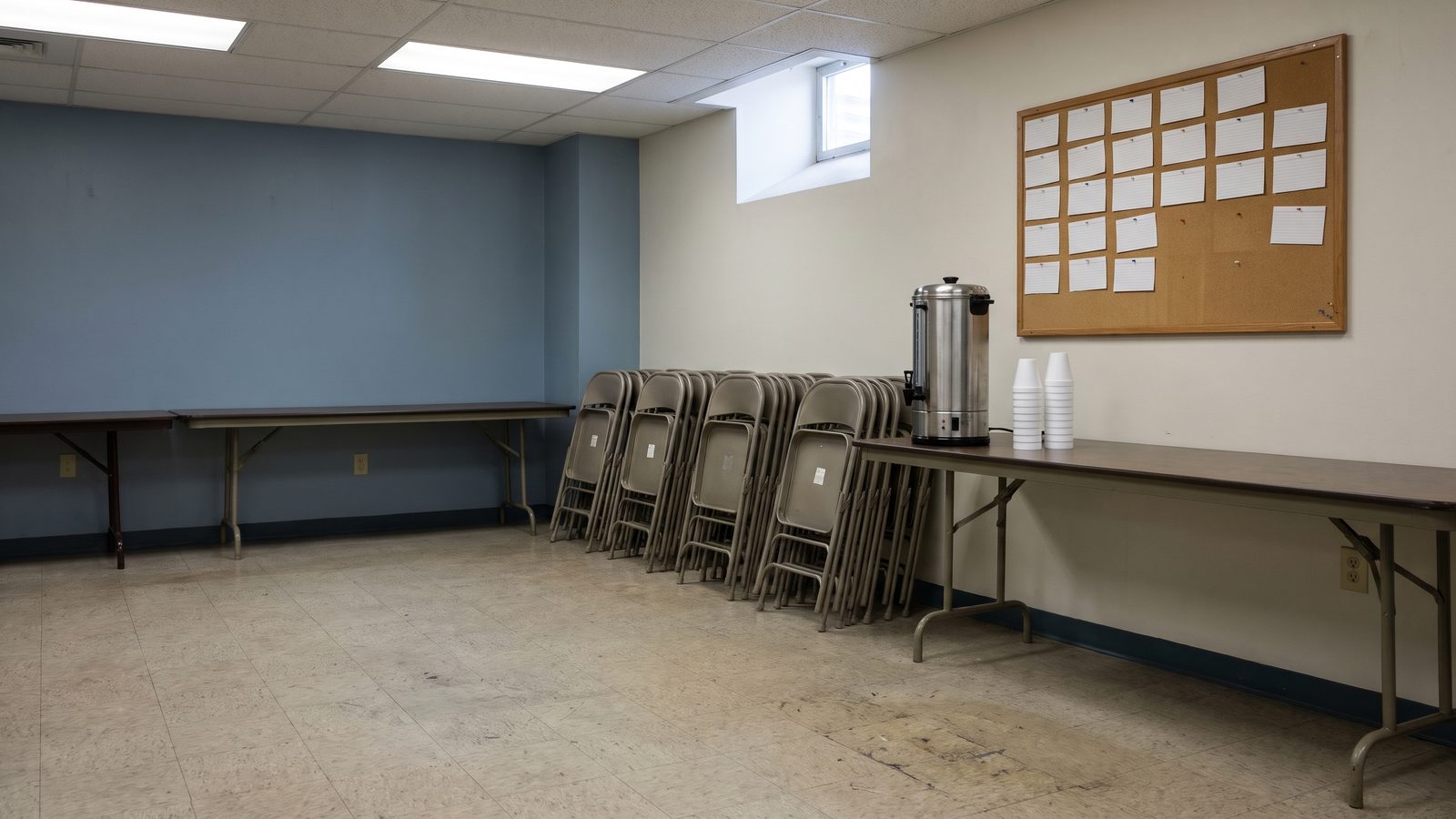 An empty basement meeting room with stacked folding chairs, a coffee urn and a cork board of blank cards