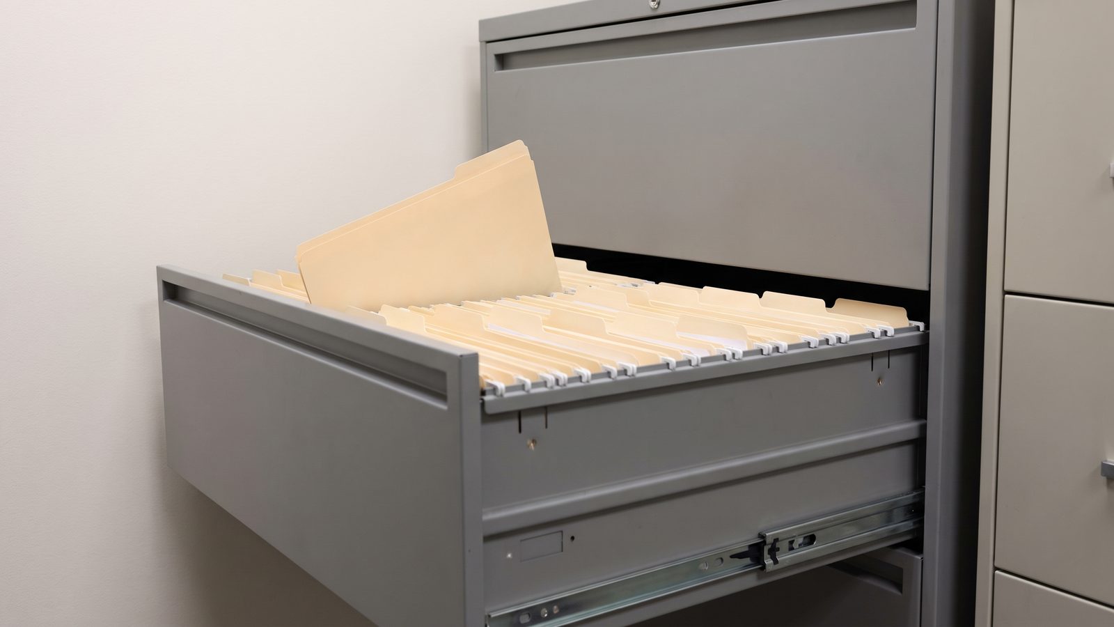 An open drawer of a gray steel personnel filing cabinet with rows of blank manila folder tabs, one folder lifted out of line.