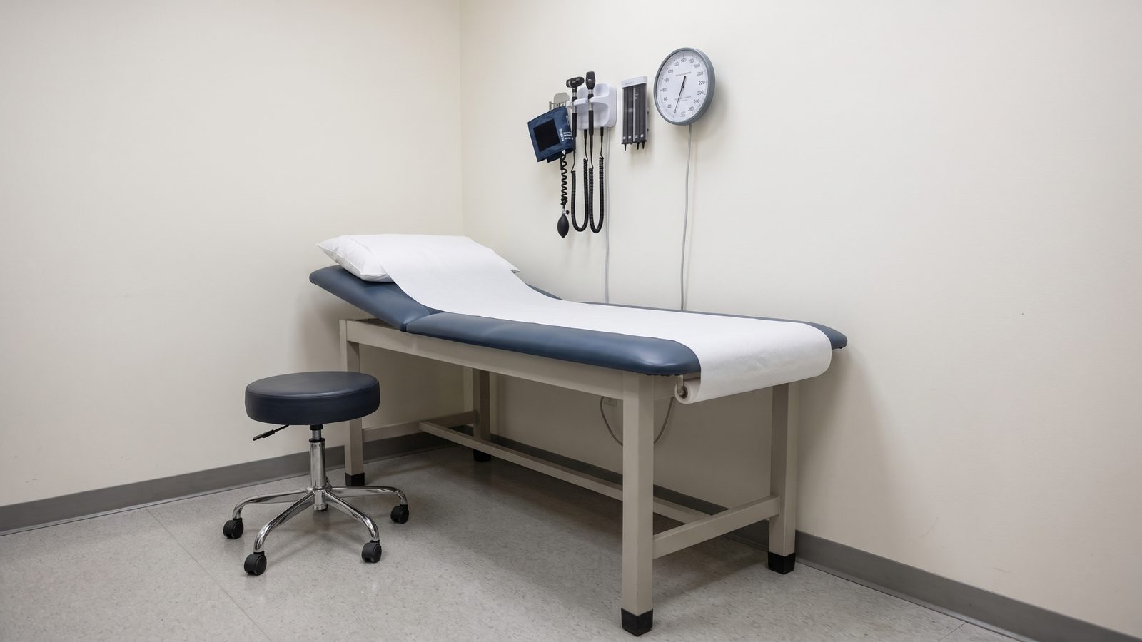 An empty medical exam room with a paper-covered exam table, a rolling stool and a wall-mounted blood pressure gauge
