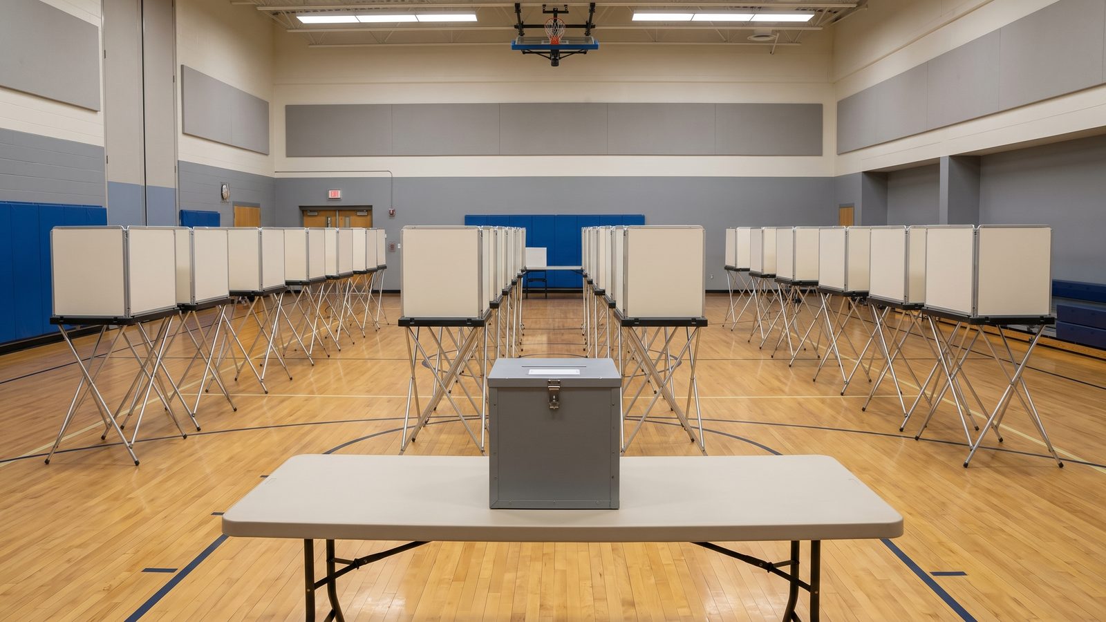 Empty school gym set up as a polling place with rows of voting booths and a gray ballot box on a folding table