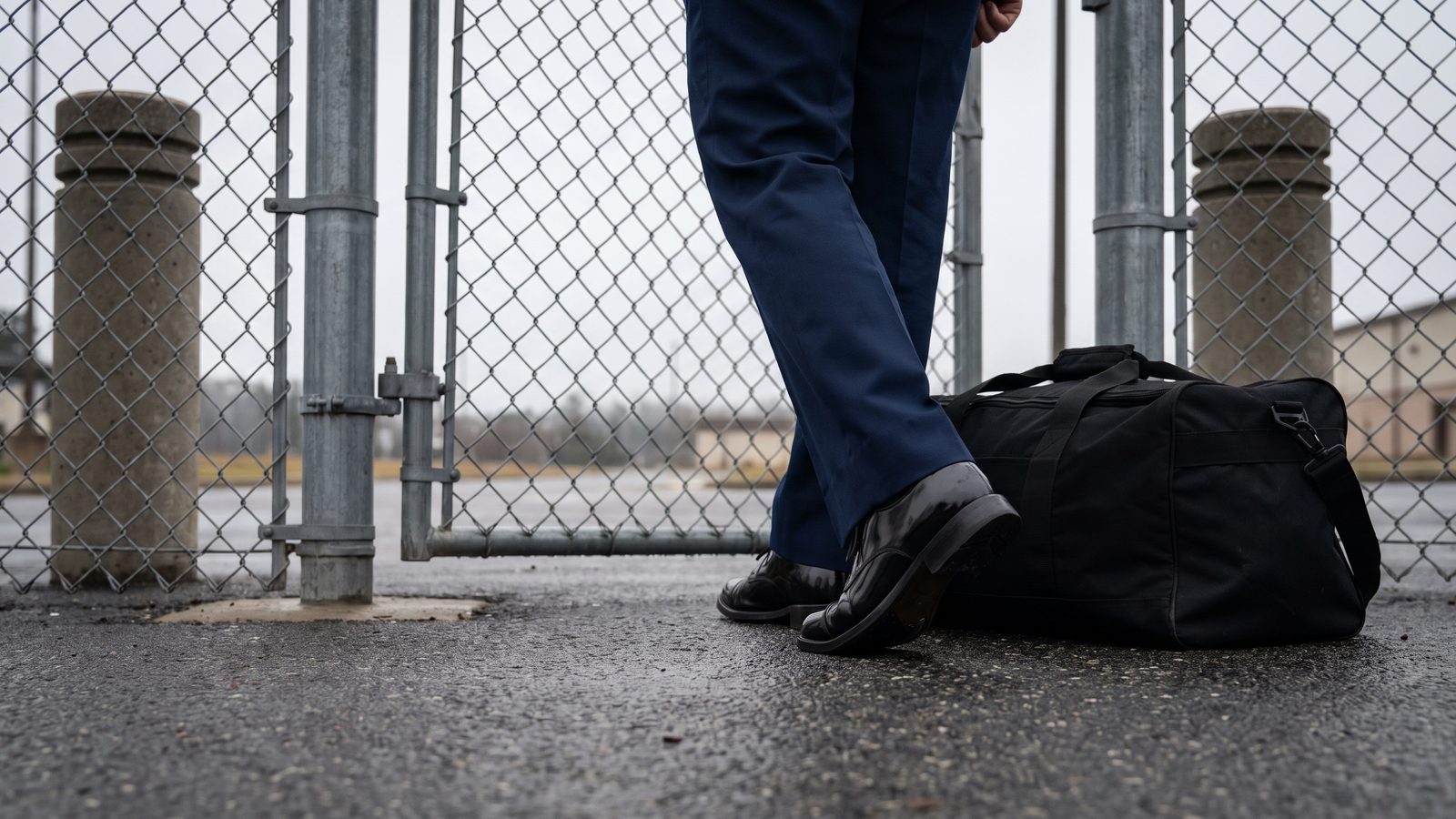 A retiring service member's dress shoes and uniform trousers at a base pedestrian gate, a black duffel bag on the wet pavement beside a chain-link fence.