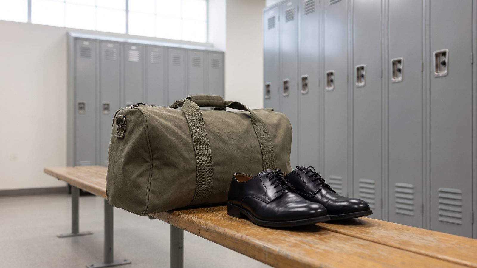 An olive canvas duffel bag and a pair of polished black dress shoes on a wooden bench in a locker room