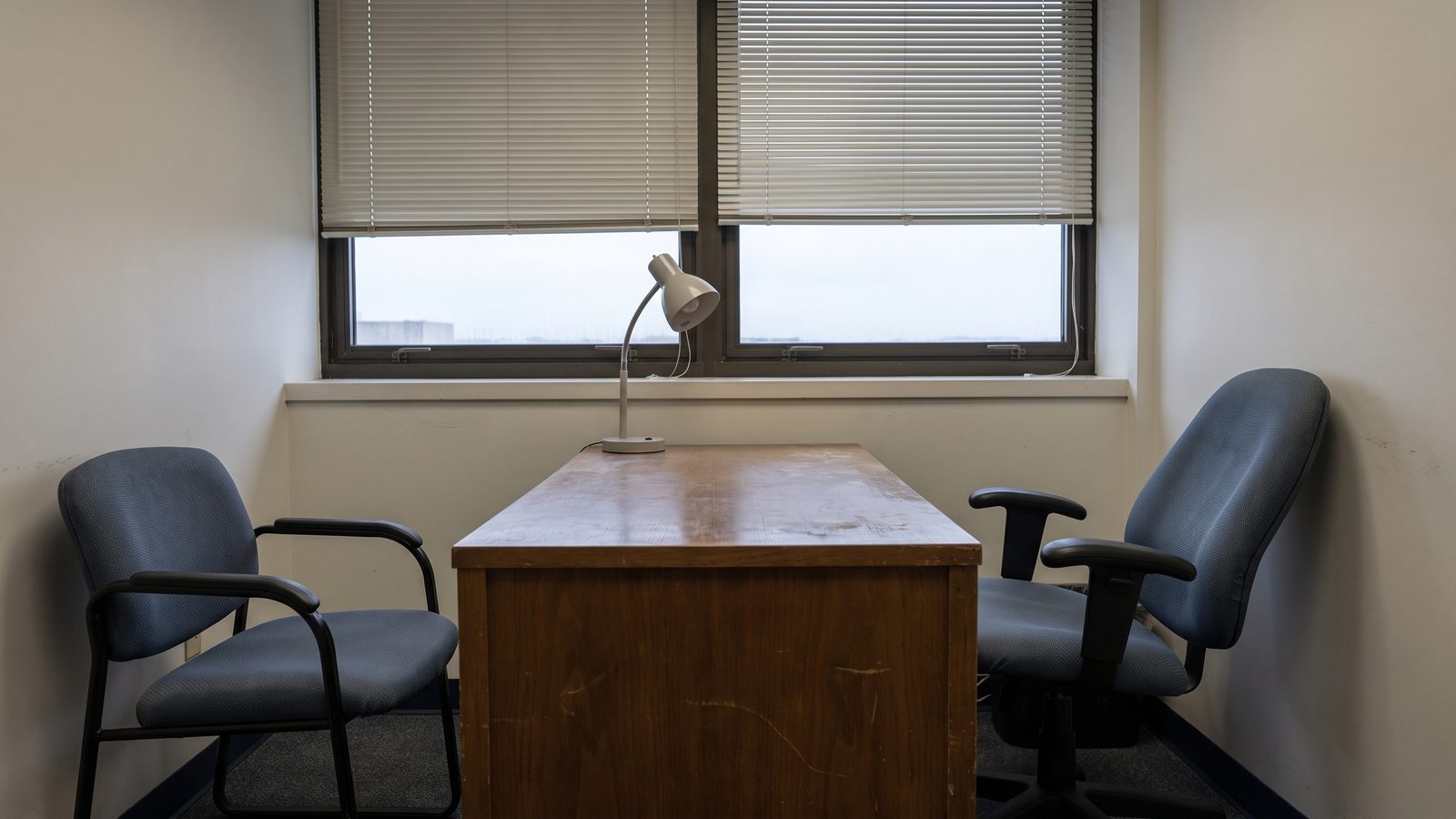 An empty federal supervisor's office at the end of the day: a bare desk with a lamp, two chairs facing each other, blinds half drawn.