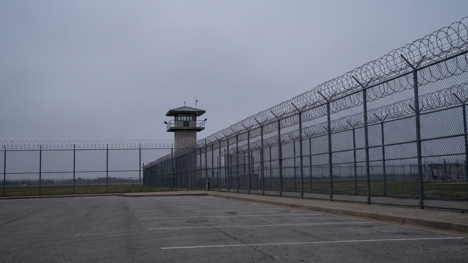 A federal prison perimeter under an overcast sky: a long chain-link fence topped with coils of razor wire, a guard tower beyond it and an empty striped parking lot in the foreground