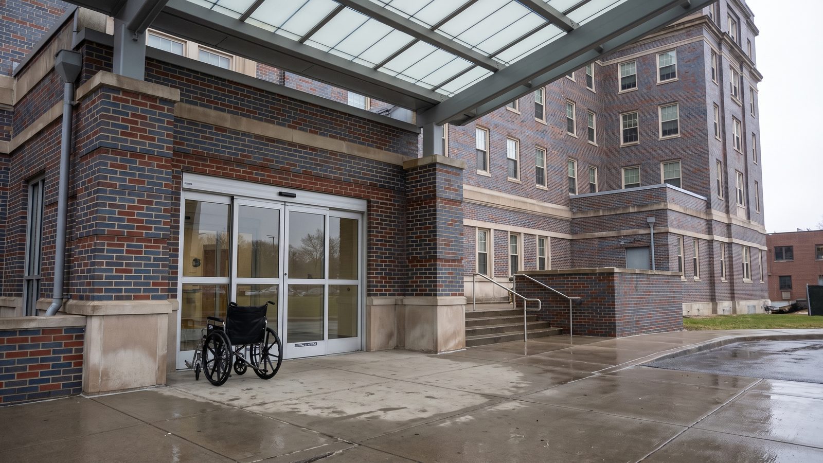 An empty wheelchair beside the glass sliding doors under the entrance canopy of a multi-story brick hospital